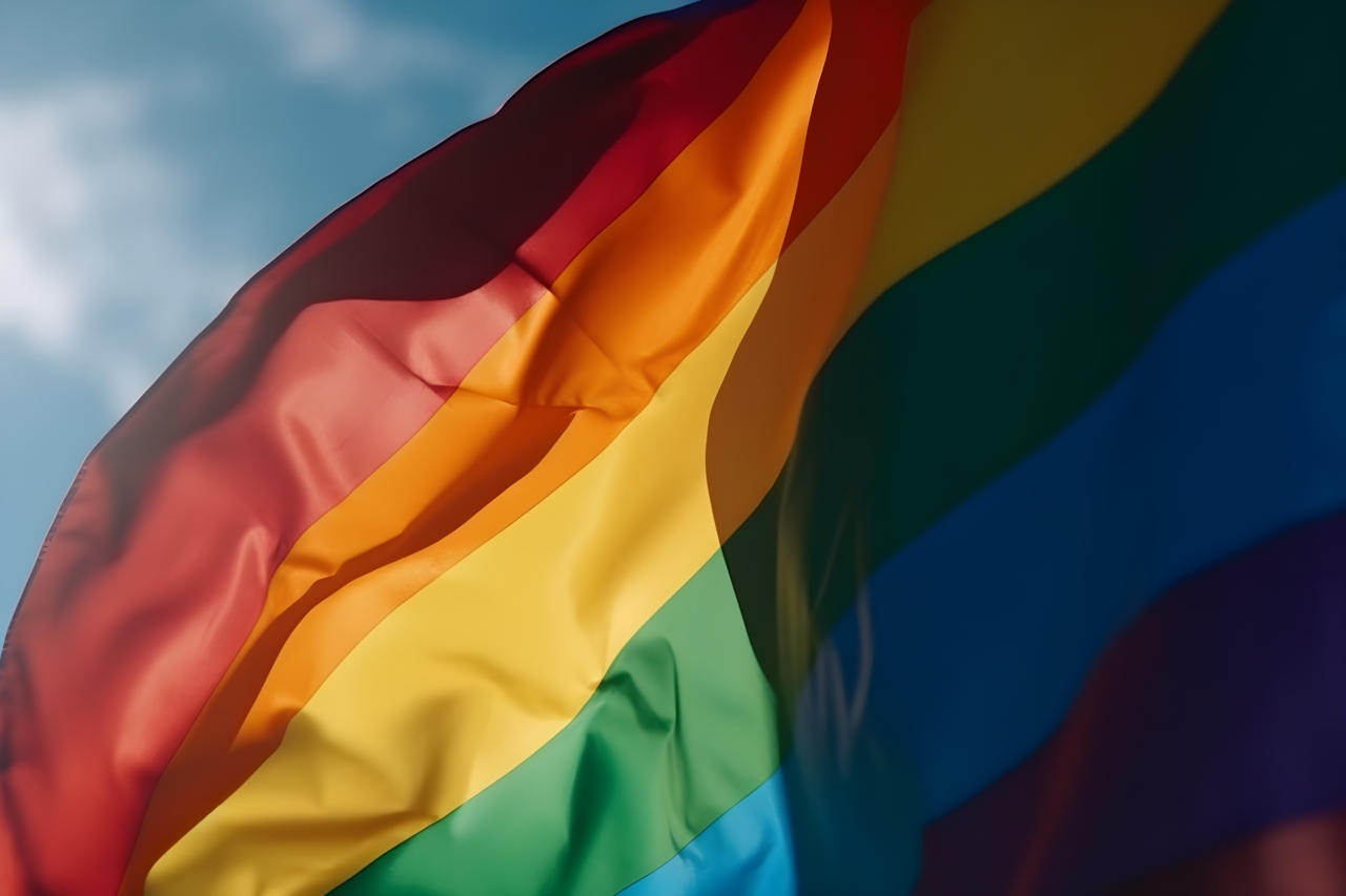 A rainbow flag waving in the wind against a blue sky with clouds