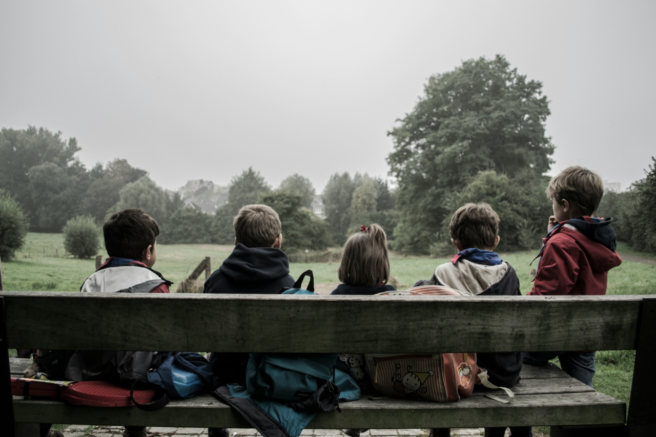 A groun of children sit on a wooden bench alongside their bags, staring off into the distance