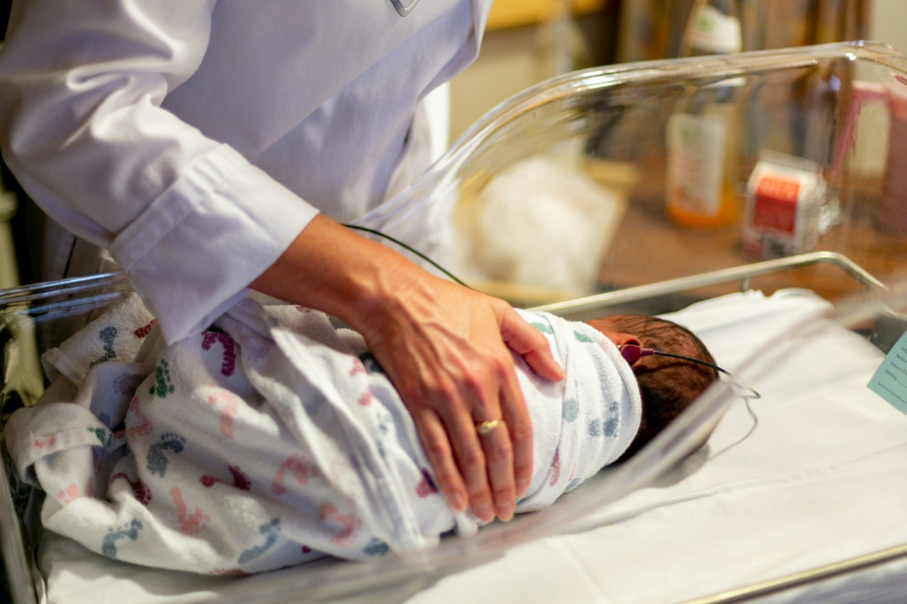 A hand lovingly cradles a swaddled newborn lying in a hospital bassinet.